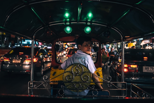 An interior view of a tuk-tuk taxi navigating through busy nighttime traffic. The driver, wearing a light blue shirt, is looking back toward the camera. Bright lights from surrounding cars illuminate the scene, reflecting off various surfaces.