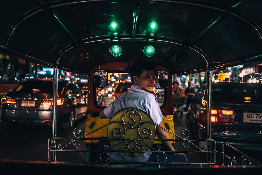 Hombre con camisa de cuello blanco sentado en autorickshaw amarillo y negro entre autos con luces traseras encendidas por la noche