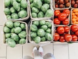 Multiple baskets are filled with fresh vegetables, including brussels sprouts and various types of tomatoes. The vegetables are neatly arranged, displaying their vibrant colors. A pair of feet wearing white shoes is visible at the bottom of the image.