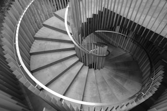 A spiral staircase viewed from above, comprised of concrete steps and a sleek metal railing. The design is modern and minimalistic, with clean lines and a symmetrical pattern. The surrounding walls are vertical slats, providing a unique architectural feature.