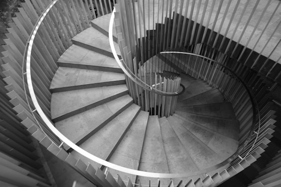 A spiral staircase viewed from above, comprised of concrete steps and a sleek metal railing. The design is modern and minimalistic, with clean lines and a symmetrical pattern. The surrounding walls are vertical slats, providing a unique architectural feature.