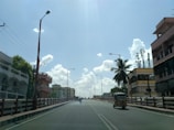 A wide road running through a city, lined with buildings on both sides. Lamp posts are evenly spaced along the road, and there are a few vehicles, including an auto rickshaw. The sky is clear with a few scattered clouds, and sunlight is bright.