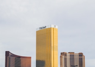 Tall skyscraper with golden reflective windows, prominently featuring the name Trump at the top. Two additional buildings are partially visible on either side, with one having a sweeping, curved facade and the other featuring an ornate, classical architecture style.
