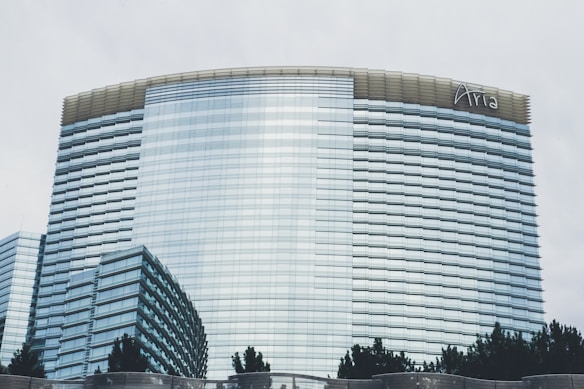 A modern high-rise building with a sleek glass facade and a curved architectural design. The structure has uniform horizontal lines and a logo at the top. Trees are visible in the foreground, and the sky appears overcast.