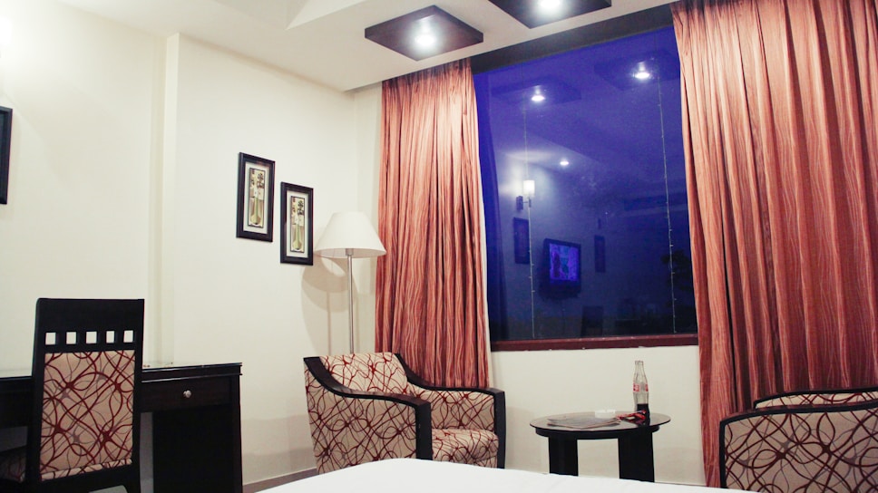 A hotel room with a contemporary design features two patterned chairs and a small round table with a bottle and magazine. The large window is framed by orange-brown curtains and displays a blue tint reflecting interior lights. Two framed artworks and a standing lamp add to the decor next to a dark desk.