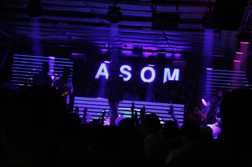 A dimly lit nightclub scene features a DJ booth illuminated by neon purple lights. The letters 'ASOM' are prominently displayed in bright white against a backdrop of horizontal light stripes. Silhouettes of people are visible, suggesting a lively party atmosphere.