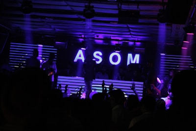 A dimly lit nightclub scene features a DJ booth illuminated by neon purple lights. The letters 'ASOM' are prominently displayed in bright white against a backdrop of horizontal light stripes. Silhouettes of people are visible, suggesting a lively party atmosphere.