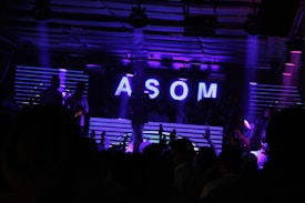 A dimly lit nightclub scene features a DJ booth illuminated by neon purple lights. The letters 'ASOM' are prominently displayed in bright white against a backdrop of horizontal light stripes. Silhouettes of people are visible, suggesting a lively party atmosphere.