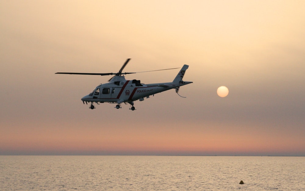 white and red helicopter flying over the sea during sunset