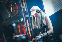 A DJ is performing in a dark, vibrant club setting. The person is focused on mixing music, wearing a white cap and headphones. The background is illuminated with bright red and orange lights, and there is high contrast between light and shadows. The DJ is operating a laptop and equipment on a table in front of them.