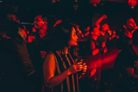 A woman is holding a drink in a dimly lit club setting, surrounded by people dancing. The ambient lighting casts a red glow over the crowd, creating a lively and energetic atmosphere.