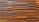 Rows of rectangular orange and black panels arranged in an alternating pattern, forming a modern and abstract facade design. The panels are mounted on a structure, creating a textured surface with shadows. A small portion of greenery is visible in the bottom left corner.