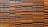 Rows of rectangular orange and black panels arranged in an alternating pattern, forming a modern and abstract facade design. The panels are mounted on a structure, creating a textured surface with shadows. A small portion of greenery is visible in the bottom left corner.