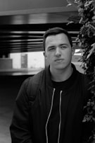 A black and white portrait of a person with short hair wearing a dark jacket and backpack, positioned in front of a structural background with overhanging beams and foliage on the side.
