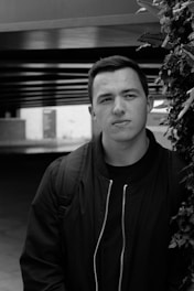 A black and white portrait of a person with short hair wearing a dark jacket and backpack, positioned in front of a structural background with overhanging beams and foliage on the side.
