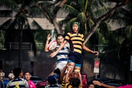 A group of rugby players are in action during a lineout. One player is lifted into the air by teammates to catch the ball. They are wearing distinct striped jerseys. Palm trees and an urban background are visible in the distance, suggesting the game is taking place outdoors in a city environment.