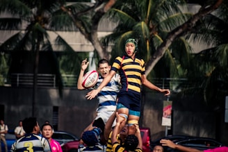 A group of rugby players are in action during a lineout. One player is lifted into the air by teammates to catch the ball. They are wearing distinct striped jerseys. Palm trees and an urban background are visible in the distance, suggesting the game is taking place outdoors in a city environment.