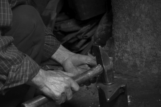A black and white photo capturing the hands of a person engaging in metalwork. The individual is holding a hammer and working on an anvil, surrounded by tools and equipment suggesting a workshop environment. The texture of the wall and the worn surfaces add a sense of age and experience.