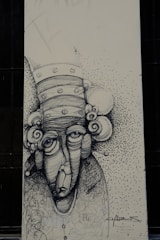 A monochromatic drawing of an abstract face with exaggerated features, including large eyes and a long nose. The head is adorned with circular elements resembling a whimsical hat or hair. The background is speckled with small dots, creating a textured effect. The artwork includes scripted text and signature at the bottom.