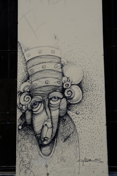 A monochromatic drawing of an abstract face with exaggerated features, including large eyes and a long nose. The head is adorned with circular elements resembling a whimsical hat or hair. The background is speckled with small dots, creating a textured effect. The artwork includes scripted text and signature at the bottom.