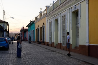 A narrow cobblestone street lined with colorful colonial-style buildings. People are casually walking and interacting along the street. The sky in the background is transitioning into sunset, adding a warm glow to the scene. The architecture features ornamental columns and wrought iron grills on the windows.