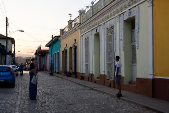 A narrow cobblestone street lined with colorful colonial-style buildings. People are casually walking and interacting along the street. The sky in the background is transitioning into sunset, adding a warm glow to the scene. The architecture features ornamental columns and wrought iron grills on the windows.