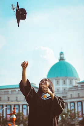A joyful graduate in a cap and gown is tossing her graduation cap into the air with a grand historical building in the background.