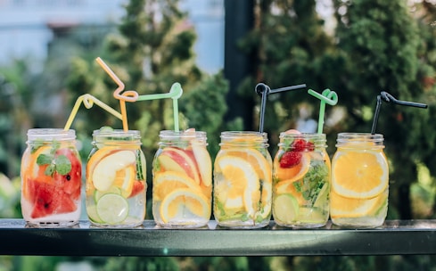 A row of five mason jars filled with infused water and colorful fruit slices is placed on a ledge. Each jar contains different combinations of fruits such as watermelon, lime, orange, lemon, and strawberries, along with mint leaves, giving a fresh and summery feel. The jars have colorful straws, and the background features lush greenery out of focus.