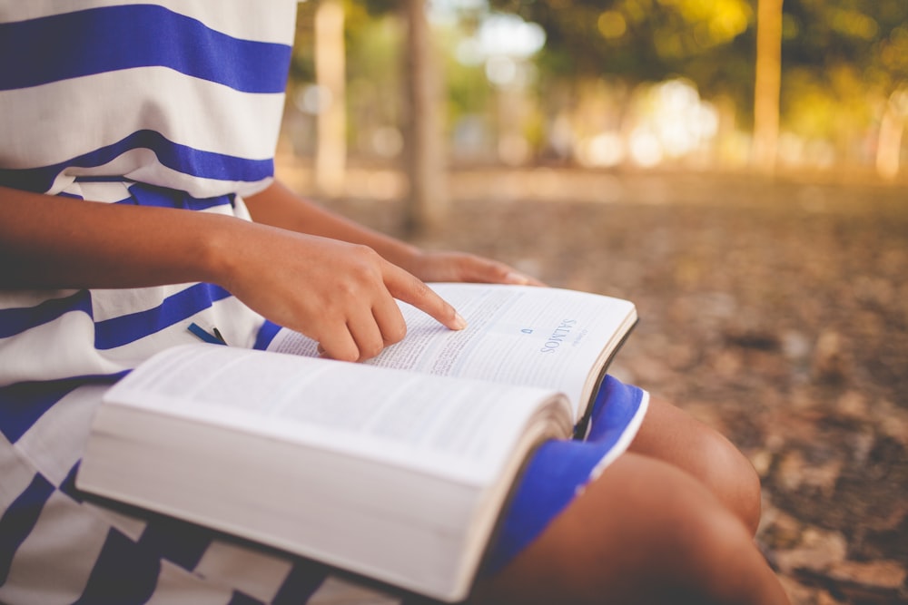 Une femme vêtue d’une robe rayée bleue et blanche assise dans un parc en train de lire son livre.