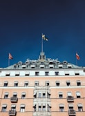 A historic hotel building with a grand and elegant facade, featuring multiple flags on its roof. The structure has a classic architectural style with many windows and balconies. The central part of the building is adorned with a tower and the hotel name is prominently displayed.