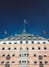 A historic hotel building with a grand and elegant facade, featuring multiple flags on its roof. The structure has a classic architectural style with many windows and balconies. The central part of the building is adorned with a tower and the hotel name is prominently displayed.