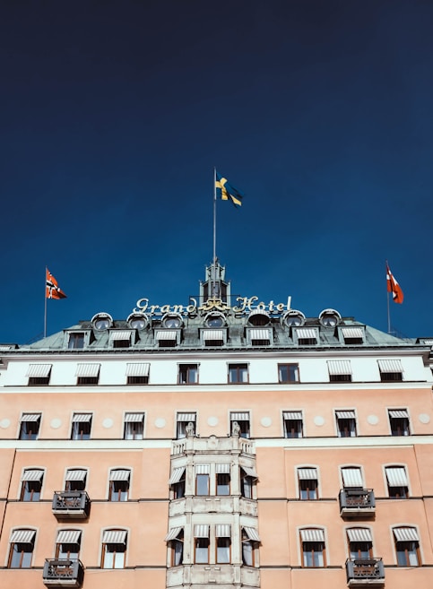 A historic hotel building with a grand and elegant facade, featuring multiple flags on its roof. The structure has a classic architectural style with many windows and balconies. The central part of the building is adorned with a tower and the hotel name is prominently displayed.