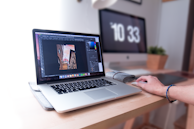 A person is using a laptop that displays a graphics editing software with a photo of buildings. The workspace includes a desktop monitor showing the time, a plant, and other office supplies on a wooden desk, suggesting a creative and organized work environment.