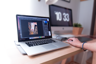 A person is using a laptop that displays a graphics editing software with a photo of buildings. The workspace includes a desktop monitor showing the time, a plant, and other office supplies on a wooden desk, suggesting a creative and organized work environment.