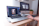A person is using a laptop that displays a graphics editing software with a photo of buildings. The workspace includes a desktop monitor showing the time, a plant, and other office supplies on a wooden desk, suggesting a creative and organized work environment.