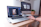 A person is using a laptop that displays a graphics editing software with a photo of buildings. The workspace includes a desktop monitor showing the time, a plant, and other office supplies on a wooden desk, suggesting a creative and organized work environment.