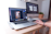 A person is using a laptop that displays a graphics editing software with a photo of buildings. The workspace includes a desktop monitor showing the time, a plant, and other office supplies on a wooden desk, suggesting a creative and organized work environment.