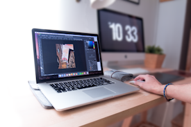 A person is using a laptop that displays a graphics editing software with a photo of buildings. The workspace includes a desktop monitor showing the time, a plant, and other office supplies on a wooden desk, suggesting a creative and organized work environment.