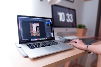 A person is using a laptop that displays a graphics editing software with a photo of buildings. The workspace includes a desktop monitor showing the time, a plant, and other office supplies on a wooden desk, suggesting a creative and organized work environment.