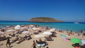 A crowded beach scene with numerous people relaxing on sunbeds and under umbrellas. The sandy shore is lined with vibrant towels and sunbathers, while people enjoy the clear blue sea. In the distance, a rocky island rises from the water, and a white yacht is anchored nearby. The sky is clear and blue, adding to the summery atmosphere.