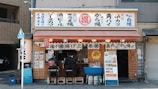A small Japanese street food stall with signage displaying various menu items. Two customers are seated on stools at the counter, which is decorated with traditional Japanese elements like lanterns and banners. A menu board with pictures of dishes is displayed on the outside.