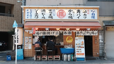 A small Japanese street food stall with signage displaying various menu items. Two customers are seated on stools at the counter, which is decorated with traditional Japanese elements like lanterns and banners. A menu board with pictures of dishes is displayed on the outside.