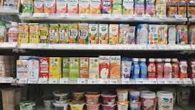 A variety of packaged drinks and yogurt products are neatly organized on grocery store shelves. The top shelf features an assortment of colorful fruit juice cartons, while the middle shelf contains more juice cartons as well as bottled drinks. The bottom shelf displays a selection of yogurt cups and beverages.