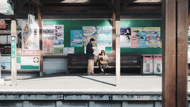 A small train station platform with two people waiting on a wooden bench. The backdrop features a variety of colorful posters and advertisements on a green bulletin board. The platform is bathed in sunlight, casting shadows on the ground, while wooden beams provide structure and support.