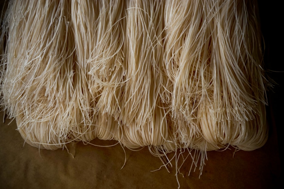 A bundle of raw, thin, light-colored noodles is arranged in parallel lines. The texture is fibrous and slightly shiny, indicating a fresh, uncooked state.