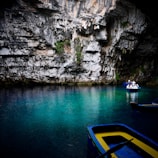 A serene underground lake with crystal clear turquoise water surrounded by rocky cave walls. Two blue and yellow wooden boats float on the water; one is empty while the other carries a group of people wearing life vests and hats. The cave walls are textured with jagged rocks and small patches of vegetation.