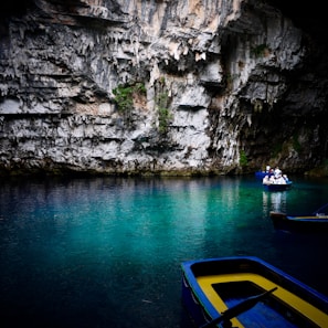 A serene underground lake with crystal clear turquoise water surrounded by rocky cave walls. Two blue and yellow wooden boats float on the water; one is empty while the other carries a group of people wearing life vests and hats. The cave walls are textured with jagged rocks and small patches of vegetation.