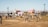 A lively outdoor festival scene with people walking and gathering in an open field. Colorful geometric structures with artistic designs are prominently displayed in the background amidst palm trees. The atmosphere is bright and sunny, indicative of a warm day.