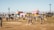 A lively outdoor festival scene with people walking and gathering in an open field. Colorful geometric structures with artistic designs are prominently displayed in the background amidst palm trees. The atmosphere is bright and sunny, indicative of a warm day.