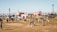 A lively outdoor festival scene with people walking and gathering in an open field. Colorful geometric structures with artistic designs are prominently displayed in the background amidst palm trees. The atmosphere is bright and sunny, indicative of a warm day.